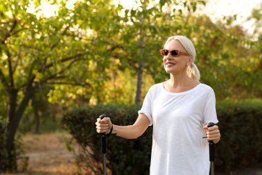 Güneş gözlüklü güzel olgun bir kadın parkta sırıklarla antrenman yapıyor.