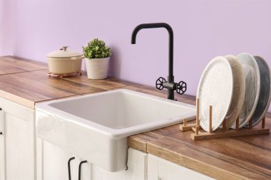 Beyaz seramik lavabo ve tabaklar mutfaktaki mor duvarın yanındaki ahşap tezgahta.