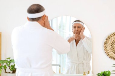 Evde aynanın yanında yüz geliştirme egzersizi yapan bornozlu olgun bir adam.