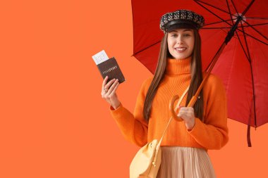 Turuncu arka planda şemsiyesi ve pasaportu olan sonbahar elbiseli bir kadın turist.