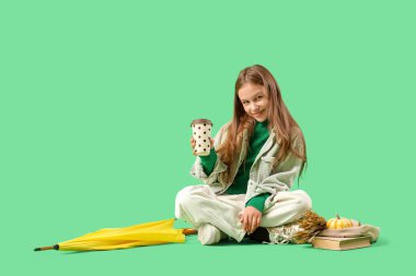 Yeşil arka planda oturan bir fincan kahve, kitap ve sarı şemsiyeli tatlı genç kız.