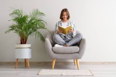 Beyaz duvarın yanındaki koltukta kitap okuyan güzel bir kadın.