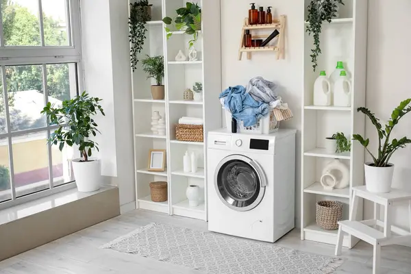 Çamaşır makinesi, çamaşır sepeti ve çiçekleri olan hafif çamaşır odasının içi.