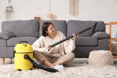 Evdeki halının üzerinde modern elektrikli süpürgeyle oturan güzel genç bir kadın.