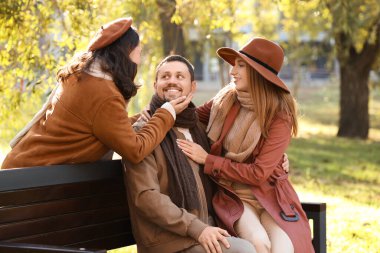 Man with two beautiful women sitting on bench in autumn park. Polyamory concept clipart