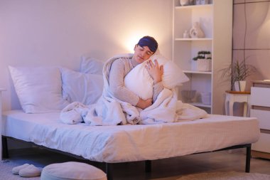 Uyku maskesi ve yastıklı uykulu genç adam evde yatakta oturuyor.