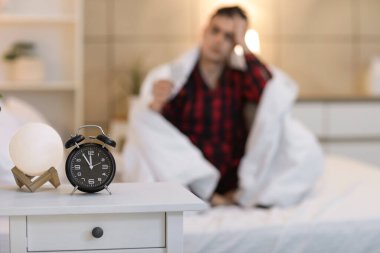 Young man with insomnia pills sitting on bed at home, focus on alarm clock clipart