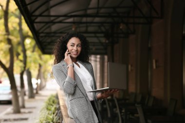 Güzel, genç, mutlu, Afro-Amerikan iş kadını. Laptopuyla sokak kafesinde cep telefonuyla konuşuyor.