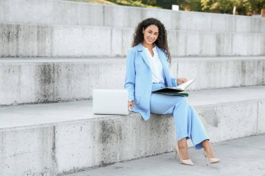 Güzel, genç, mutlu, Afro-Amerikan iş kadını dizüstü bilgisayarı ve belgeleriyle şehirdeki merdivenlerde oturuyor.