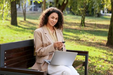 Bilgisayarı ve cep telefonuyla parkta bankta oturan güzel, mutlu, Afro-Amerikan iş kadını.