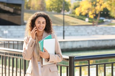 Dizüstü bilgisayarı ve klasörü olan güzel, genç, mutlu, Afro-Amerikan iş kadını bariyer kenarında cep telefonuyla konuşuyor.