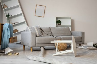 Kanepe, raf üniteleri ve deprem sonrası devrilen masalarla dolu dağınık bir oturma odası.