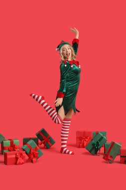 Kırmızı arka planda Noel hediyeleri olan elf gibi giyinmiş mutlu genç bir kadın.