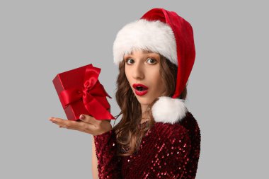Noel Baba şapkalı, gri arka planda Noel hediyesi kutusu olan şaşırmış genç bir kadın.