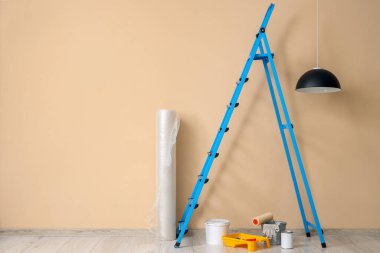 Kabarcık ambalajlı merdiven, boya kutuları ve bej duvarın yanındaki aletler.