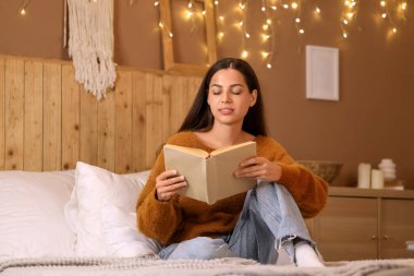 Güzel, genç, mutlu bir kadın akşamları yatakta kitap okuyor.