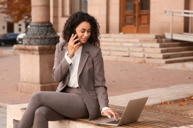 Güzel, genç, mutlu, Afro-Amerikan iş kadını. Laptopuyla şehirdeki bankta cep telefonuyla konuşuyor.