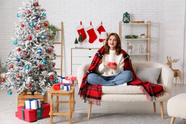 Bir fincan sıcak kakaolu güzel bir kadın Noel için dekore edilmiş bir odada koltukta oturuyor.