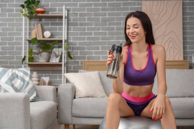 Evde spor salonunda su şişesiyle oturan genç bir kadın.