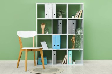 Ofisteki yeşil duvarın yanında doküman klasörleri, bitkileri ve sandalyesi olan raf ünitesi