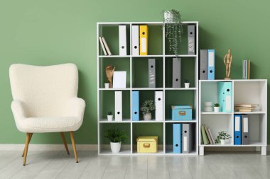 Ofiste yeşil duvarın yanında doküman klasörleri, bitkileri ve koltuğu olan raflar