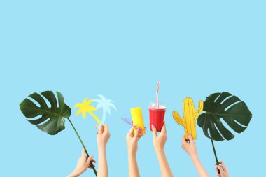 Kokteyl, güneş kremi, gözlük, canavar yaprakları, kağıt kaktüs ve mavi arka planda palmiye ağaçları olan kadın elleri. Seyahat kavramı