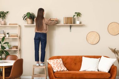 Güzel genç bir kadın oturma odasındaki raftan kitap alıyor.