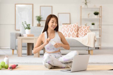 Evde yoga minderinde laptoplu ve lezzetli yoğurtlu genç Asyalı kadın.