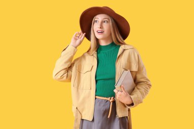 Sarı arka planda kitabı olan şık şapkalı güzel bir genç kadın.