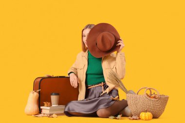 Elinde kahve, bavul, kitap, balkabağı ve sonbahar yapraklarıyla sarı arka planda oturan güzel bir kadın.