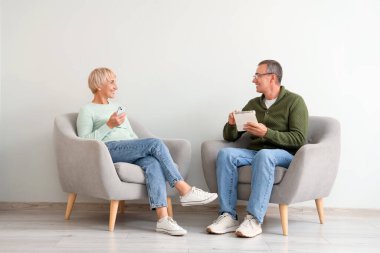 Not defteri ve cep telefonu olan yaşlı çift beyaz duvarın yanındaki koltuklarda oturuyor.