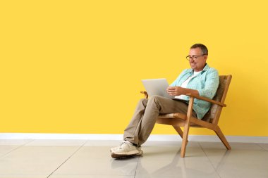 Dizüstü bilgisayarı olan yaşlı adam sarı duvarın yanında oturuyor.