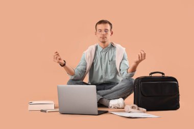 Gözlüklü genç işadamı dizüstü bilgisayarı ve çantasıyla bej arka plan üzerinde meditasyon yapıyor.
