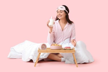 Pembe arka planda battaniyeli çizgili pijamalı genç bir kadın kahvaltı yapıyor.