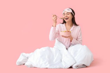 Çizgili pijamalı ve battaniyeli genç bir kadın pembe arka planda gevrek halkaları yiyor.