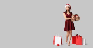 Noel Baba şapkalı güzel bir kadın. Bir sürü alışveriş çantası ve gri arka planda Noel hediyeleri var. Mesaj için yeri var.