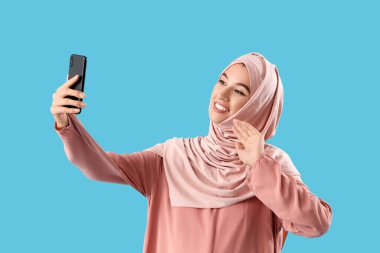 Cep telefonlu güzel Müslüman kadın mavi arka planda selfie çekiyor.