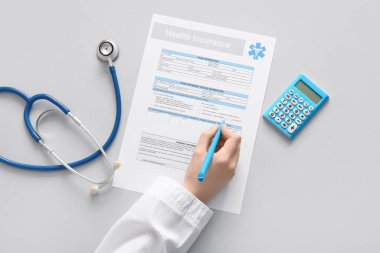 Beyaz arka planda steteskop ve hesap makinesinin yanında sağlık sigortası formu dolduran doktor.