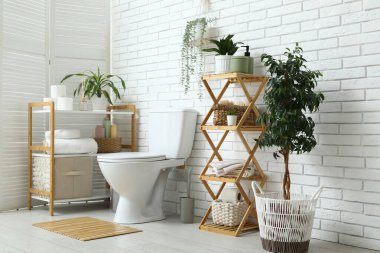 Beyaz tuğla duvarın yanında seramik klozet, ev bitkisi ve raf ünitesi olan modern tuvalet.