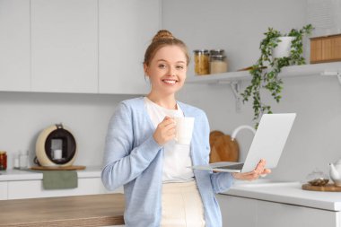 Mutfakta bir fincan kahve ve modern dizüstü bilgisayarı olan genç bir kadın.