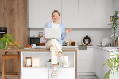 Genç kadın mutfaktaki masada modern dizüstü bilgisayar kullanıyor.