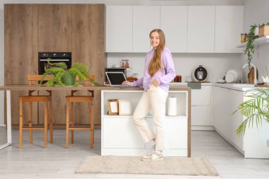 Genç kadın mutfaktaki masada modern dizüstü bilgisayar kullanıyor.