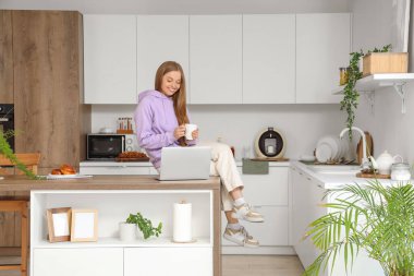 Bir fincan kahve ve modern dizüstü bilgisayarı olan genç bir kadın mutfaktaki masada oturuyor.