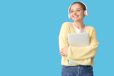 Mavi arka planda modern dizüstü bilgisayarı olan kulaklıklı genç bir kadın.