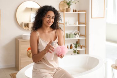 Duş jeli ve banyo süngeri şişesiyle evde oturan güzel, mutlu, Afro-Amerikan kadın.