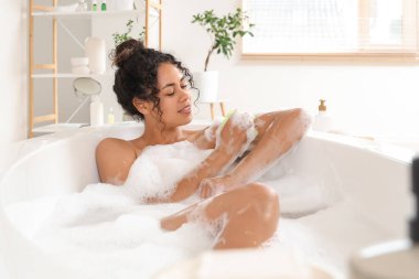 Güzel, genç, mutlu, Afro-Amerikan bir kadın evde sünger ve sabun köpüğüyle banyo yapıyor.