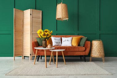 Yeşil duvarın yanındaki sehpanın üzerinde turuncu kanepesi ve vazo vazosu olan şık bir oturma odası.