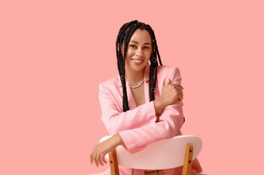 Saçları örgülü, pembe arka planda oturan seksi Afro-Amerikan kadın.