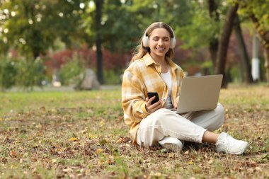 Sonbahar günü park yerinde dizüstü bilgisayar ve cep telefonu kullanan kulaklıklı bir kadın turist