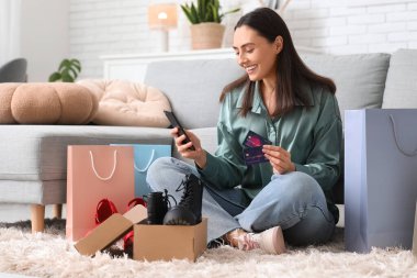 Cep telefonu, kredi kartı ve alışveriş çantalarıyla evde oturan mutlu genç bir kadın. Kara Cuma indirimi